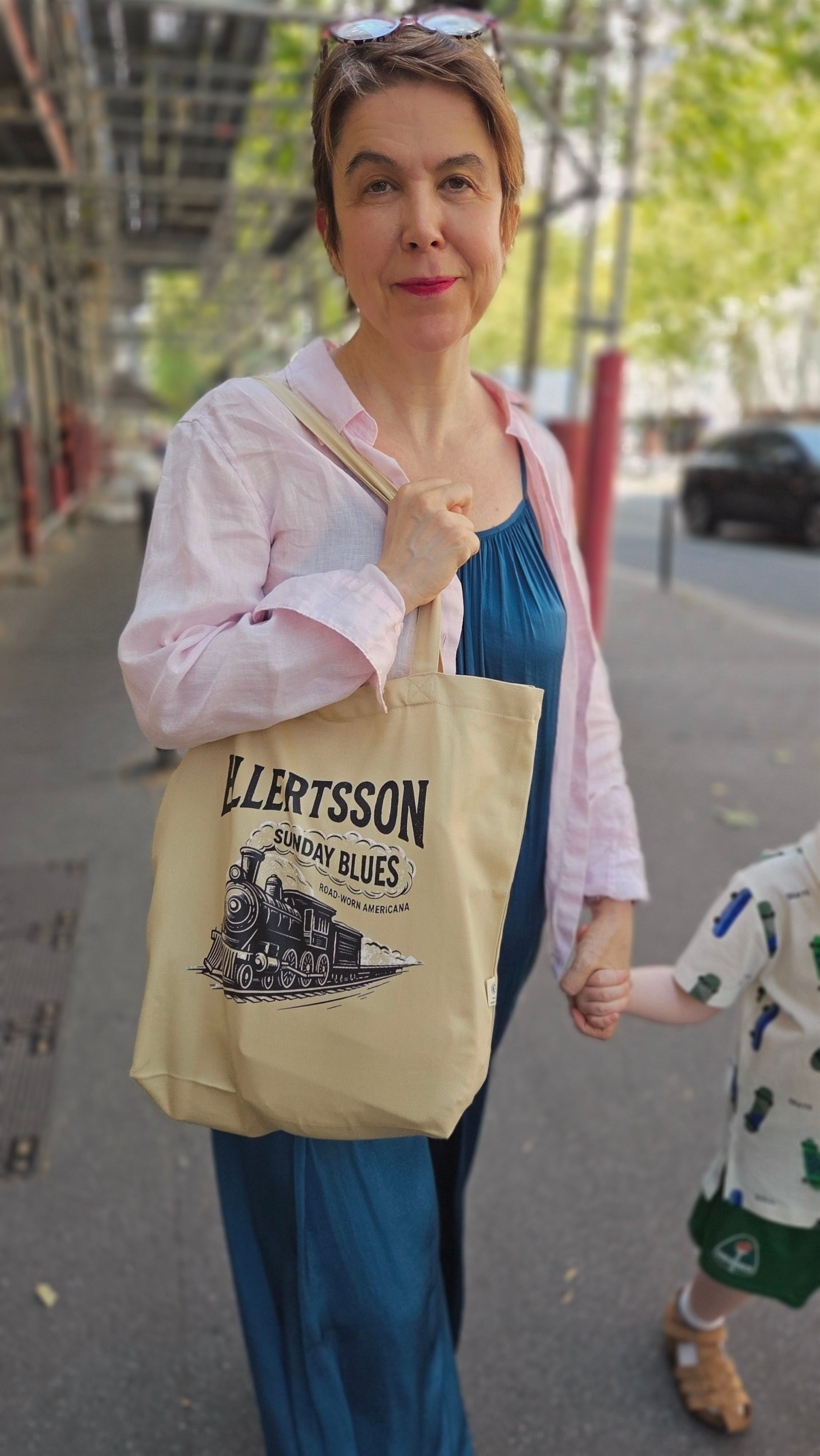 A woman carrying a beige tote bag with 'ELLRTSSON RAINY DAYS' printed on it, walking outdoors on a city street while holding a child's hand.
